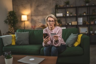 Olgun beyaz kadın kredi kartını evde online alışveriş için tutuyor.