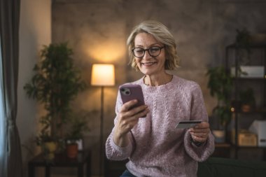 Olgun beyaz kadın kredi kartını evde online alışveriş için tutuyor.