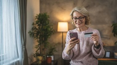 Olgun beyaz kadın kredi kartını evde online alışveriş için tutuyor.