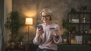 Olgun beyaz kadın kredi kartını evde online alışveriş için tutuyor.