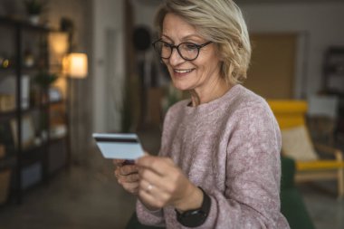 Olgun beyaz kadın kredi kartını evde online alışveriş için tutuyor.