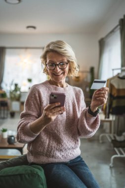 Olgun beyaz kadın kredi kartını evde online alışveriş için tutuyor.