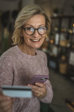 Olgun beyaz kadın kredi kartını evde online alışveriş için tutuyor.