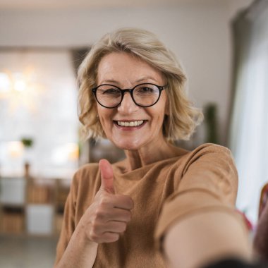 Evde gözlüklü, olgun, beyaz bir kadının portresi.