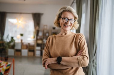 Evde gözlüklü, olgun, beyaz bir kadının portresi.