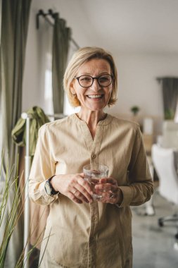 Yetişkin bir kadının portresi. Evde bir bardak su tutuyor.