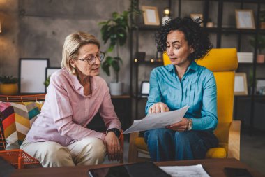 İki yetişkin kadın, ellerinde kağıt tutarak evde oturuyorlar. Evrak tutuyorlar. Danışman iş kadını, gerçek iş ve sorunlar hakkında konuşuyor.