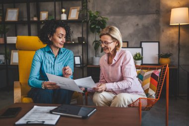 İki yetişkin kadın, ellerinde kağıt tutarak evde oturuyorlar. Evrak tutuyorlar. Danışman iş kadını, gerçek iş ve sorunlar hakkında konuşuyor.