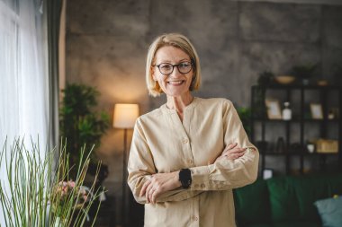 Evde gözlüklü, olgun, beyaz bir kadının portresi.