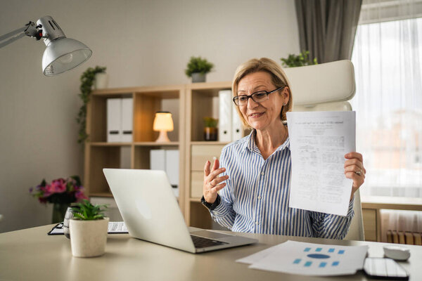 зрелая женщина-блондинка бизнес есть видео-звонок использовать ноутбук в офисе для онлайн-консультации или конференции показать бумажные документы контракт предложение или статистика