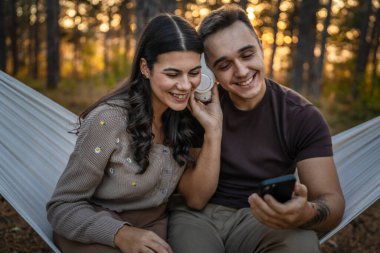 Doğadaki genç erkek ve kadın yetişkin çift kulaklıkla müzik dinliyor cep telefonu kullanıyor ya da video arama uygulaması yapıyor.