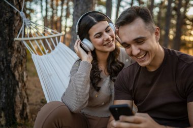 Doğadaki genç erkek ve kadın yetişkin çift kulaklıkla müzik dinliyor cep telefonu kullanıyor ya da video arama uygulaması yapıyor.
