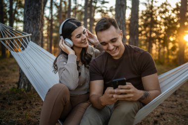 Doğadaki genç erkek ve kadın yetişkin çift kulaklıkla müzik dinliyor cep telefonu kullanıyor ya da video arama uygulaması yapıyor.