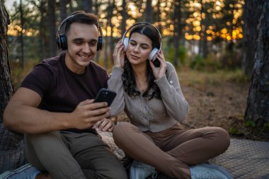 Doğadaki genç erkek ve kadın yetişkin çift kulaklıkla müzik dinliyor cep telefonu kullanıyor ya da video arama uygulaması yapıyor.
