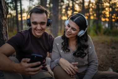 Doğadaki genç erkek ve kadın yetişkin çift kulaklıkla müzik dinliyor cep telefonu kullanıyor ya da video arama uygulaması yapıyor.