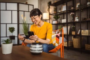 Gözlüklü Japon kadın sandalyeye oturur kredi kartı ve cep telefonunu evde online alışveriş yaparak kullanır.