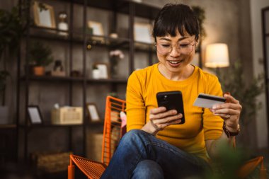 Gözlüklü Japon kadın sandalyeye oturur kredi kartı ve cep telefonunu evde online alışveriş yaparak kullanır.