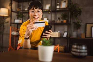 Gözlüklü Japon kadın sandalyeye oturur kredi kartı ve cep telefonunu evde online alışveriş yaparak kullanır.