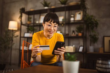 Gözlüklü Japon kadın sandalyeye oturur kredi kartı ve cep telefonunu evde online alışveriş yaparak kullanır.