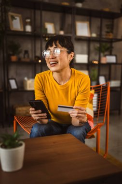 Gözlüklü Japon kadın sandalyeye oturur kredi kartı ve cep telefonunu evde online alışveriş yaparak kullanır.