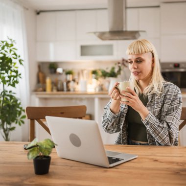 Genç, beyaz, sarışın kadın dizüstü bilgisayarda çalışıyor ve elinde bardakla evden mutlu bir pozitifti.