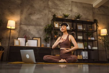 Japon bir kadın evde yoga pratiği yapıyor. İnternette meditasyon yapıyor. Kulaklıkları başında, dizüstü bilgisayar kullanıyor.