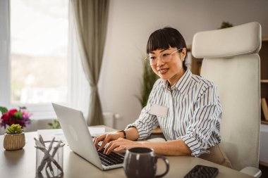 Gözlüklü olgun Japon kadın ofiste dizüstü bilgisayarla çalışıyor.