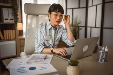 Gözlüklü olgun Japon kadın ofiste dizüstü bilgisayarla çalışıyor.