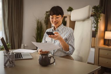 Gözlüklü, olgun Japon iş kadını. Ofis gülüşünde oturup evrakları tara, mutlu ol.