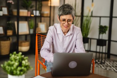 Gözlüklü olgun beyaz kadın dizüstü bilgisayardan gülümseyerek evde çalışıyor.
