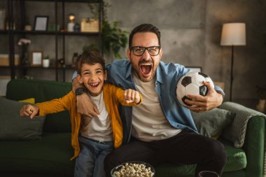 Baba ve oğlu Kafkas futbol maçı izleyip evde tezahürat yapıyorlar. Gürültülü, heyecanlı, mutlu.