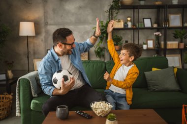 Baba ve oğlu Kafkas futbol maçı izleyip evde tezahürat yapıyorlar. Gürültülü, heyecanlı, mutlu.