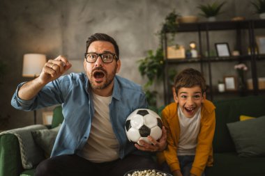 Baba ve oğlu Kafkas futbol maçı izleyip evde tezahürat yapıyorlar. Gürültülü, heyecanlı, mutlu.