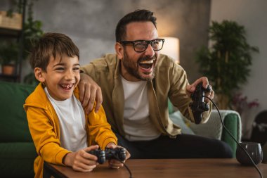 Baba ve oğlu beyaz tenli, joystick ile video oyunu oynuyor ve evde eğleniyor.