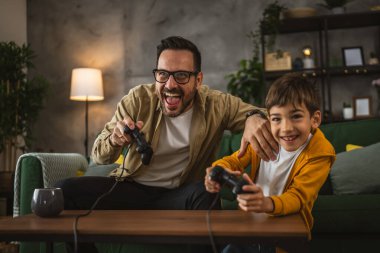 Baba ve oğlu beyaz tenli, joystick ile video oyunu oynuyor ve evde eğleniyor.