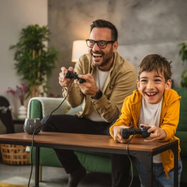 Baba ve oğlu beyaz tenli, joystick ile video oyunu oynuyor ve evde eğleniyor.