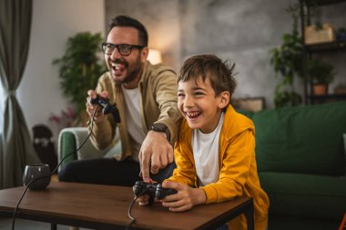 Baba ve oğlu beyaz tenli, joystick ile video oyunu oynuyor ve evde eğleniyor.