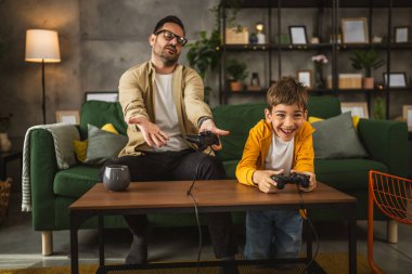 Baba ve oğlu beyaz tenli, joystick ile video oyunu oynuyor ve evde eğleniyor.