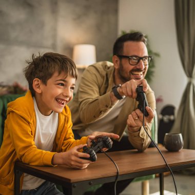 Baba ve oğlu beyaz tenli, joystick ile video oyunu oynuyor ve evde eğleniyor.