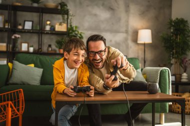 Baba ve oğlu beyaz tenli, joystick ile video oyunu oynuyor ve evde eğleniyor.