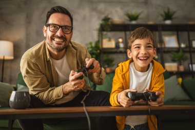 Baba ve oğlu beyaz tenli, joystick ile video oyunu oynuyor ve evde eğleniyor.