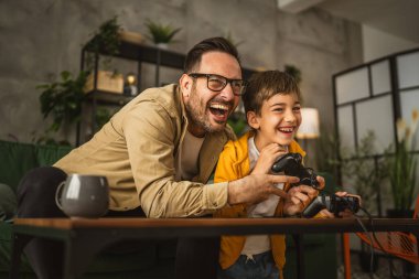 Baba ve oğlu beyaz tenli, joystick ile video oyunu oynuyor ve evde eğleniyor.