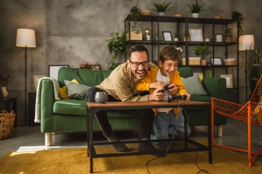 Baba ve oğlu beyaz tenli, joystick ile video oyunu oynuyor ve evde eğleniyor.