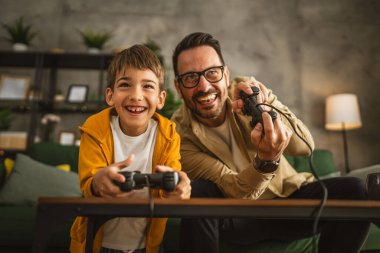 Baba ve oğlu beyaz tenli, joystick ile video oyunu oynuyor ve evde eğleniyor.