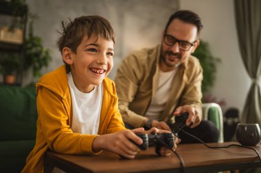 Baba ve oğlu beyaz tenli, joystick ile video oyunu oynuyor ve evde eğleniyor.