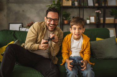 Baba ve oğlu beyaz tenli, joystick ile video oyunu oynuyor ve evde eğleniyor.