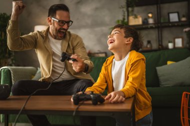 Baba ve oğlu beyaz tenli, joystick ile video oyunu oynuyor ve evde eğleniyor.