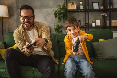 Baba ve oğlu beyaz tenli, joystick ile video oyunu oynuyor ve evde eğleniyor.