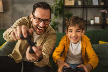 Baba ve oğlu beyaz tenli, joystick ile video oyunu oynuyor ve evde eğleniyor.
