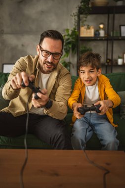 Baba ve oğlu beyaz tenli, joystick ile video oyunu oynuyor ve evde eğleniyor.